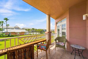 A balcony with a wooden table and chairs, a wicker chair with a cushion, and a small round table. - Seaside at Anastasia A201 – Salt, Sand & Sidewalk Cafés (St. Augustine)