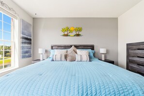 Quarto com cama grande, duas mesas de cabeceira, um criado-mudo, janela com vista e uma obra de arte na parede.