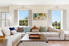 A living room with a sectional sofa, a coffee table, and a painting of palm trees. - Seaside at Anastasia A201 – Salt, Sand & Sidewalk Cafés (St. Augustine)