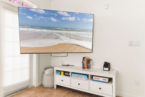 A television mounted on a wall displaying a beach scene. Below the TV, there is a white entertainment center with books and a remote control. - Seaside at Anastasia A201 – Salt, Sand & Sidewalk Cafés (St. Augustine)