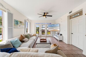 A spacious living room with a ceiling fan, a large TV, and a view of the outdoors through the glass doors. - Seaside at Anastasia A201 – Salt, Sand & Sidewalk Cafés (St. Augustine)