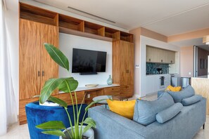 A modern living room with a flat-screen TV, wooden cabinetry, a blue sofa, and a kitchen area. - Largest Pool in Zona Romantica at SOHO PV (Puerto Vallarta)