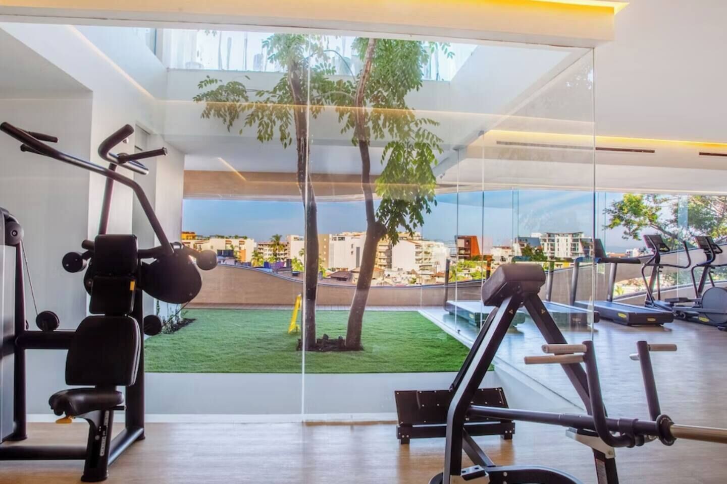 Een sportschool met trainingsapparatuur, een gebied met kunstgras en uitzicht op gebouwen door grote ramen.