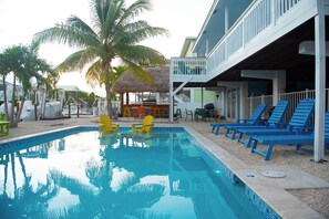 Ein Swimmingpool mit Liegestühlen und einem Strohdachschirm, umgeben von Palmen und einem Gebäude mit Balkon.