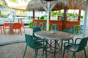 An outdoor seating area with a round table and multiple chairs, a thatched roof structure, and a wooden fence. - Gulf-to-Atlantic Waterfront Escape — Pool, Tiki Bar & Endless Fun (Marathon)