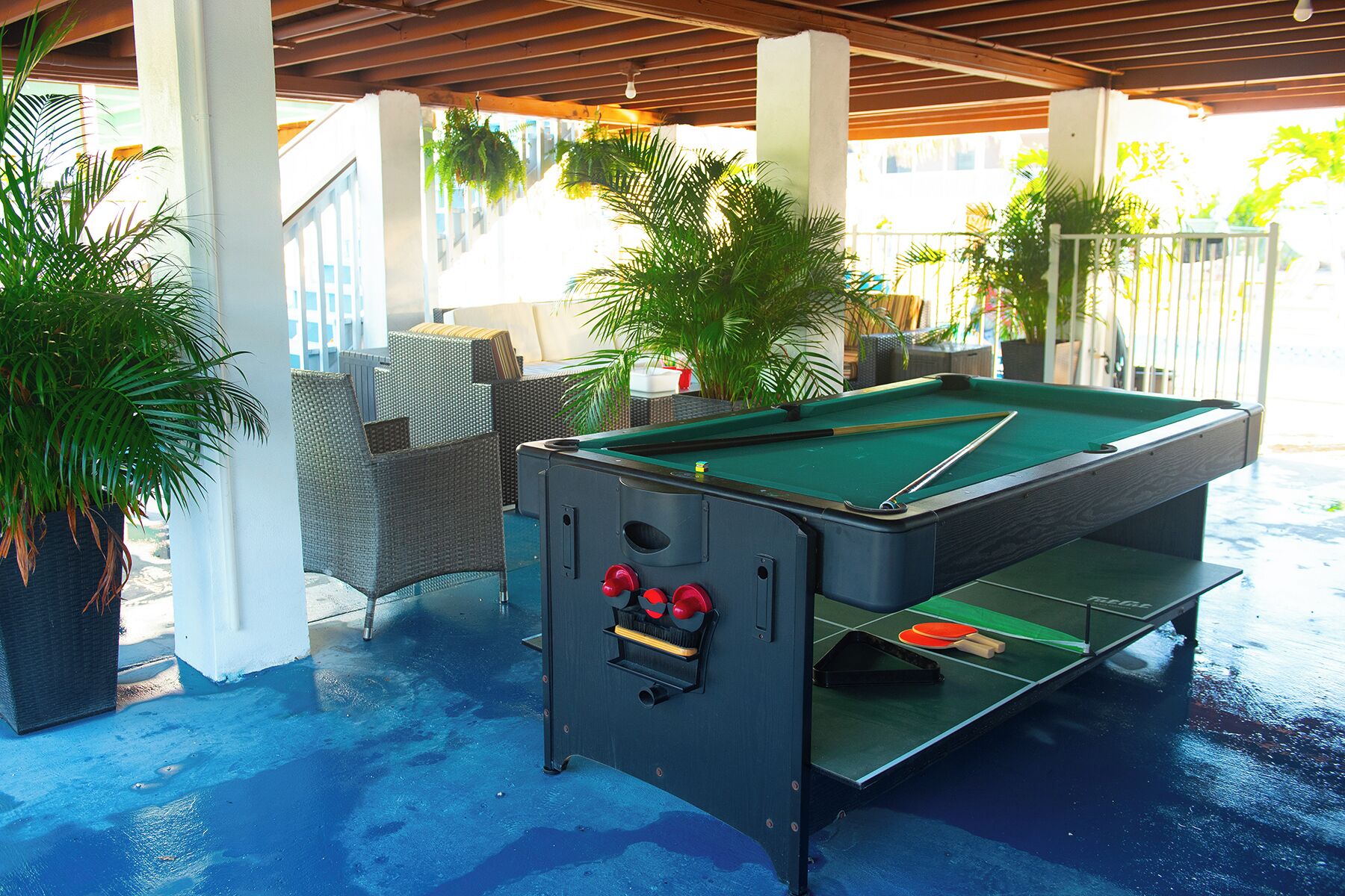 A pool table with a cue stick, surrounded by wicker furniture and potted plants in a covered outdoor area.