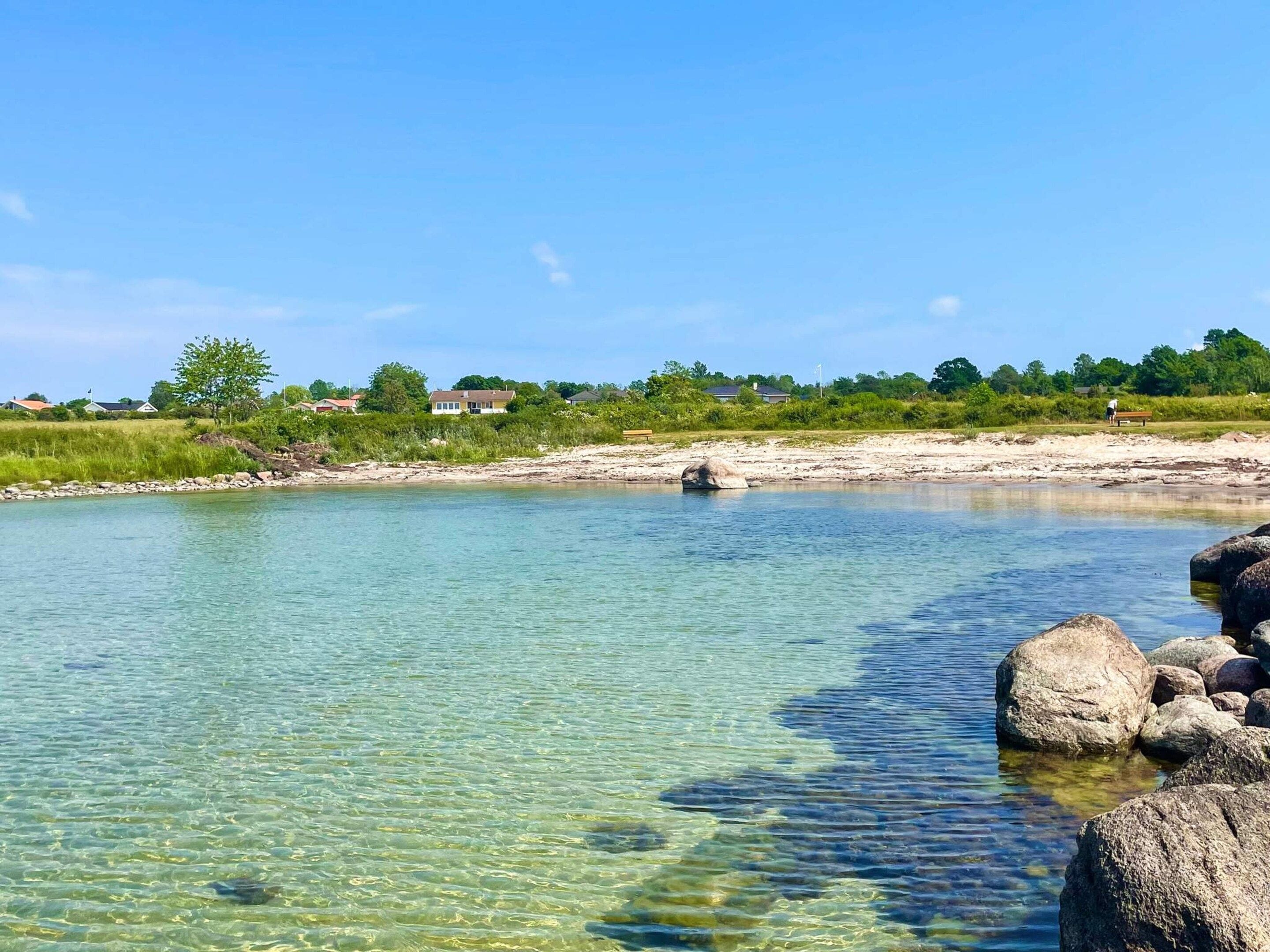 A clear river with rocks and a distant shoreline with houses.