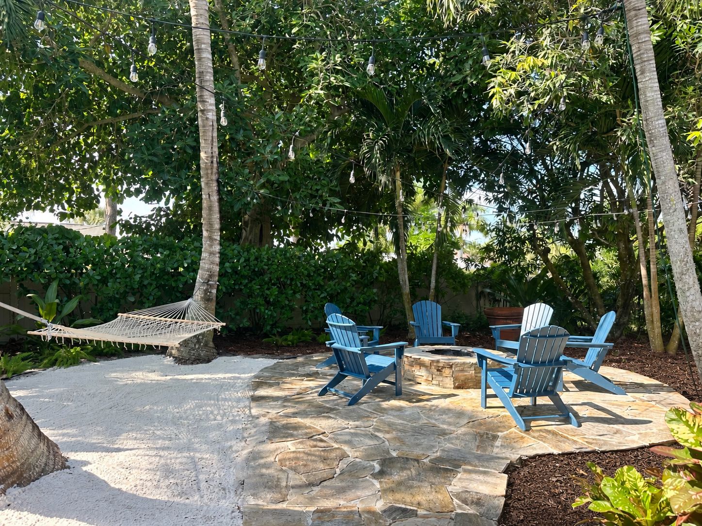 A patio with stone flooring, a hammock, and blue Adirondack chairs around a fire pit.