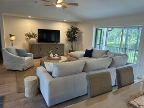 A modern living room with a sectional sofa, a television, and a large window with a view of greenery.