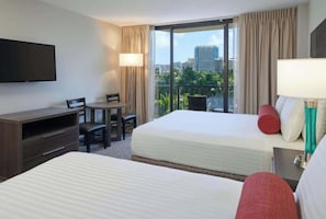 Ein Hotelzimmer mit zwei Betten, einem Schreibtisch, einem Stuhl, einem Fernseher und einem Fenster mit Blick auf die Stadt.