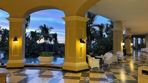 Een hotel lobby met een zwart-wit schaakbordvloer, gele zuilen en uitzicht op palmbomen en een zonsondergang.
