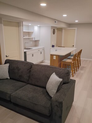 Un salon moderne avec un canapé gris foncé, une table à manger en bois avec des chaises, et un coin cuisine doté de meubles blancs.