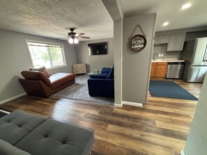 A spacious living area with a sofa, a blue chair, and a grey ottoman. A ceiling fan and a flat-screen TV are mounted on the wall. - Boji Park Shuffle: 5-bedroom home in the middle of all the action (Arnolds Park)