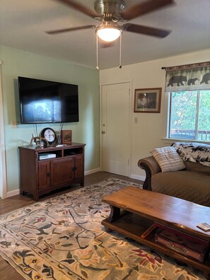 A living room with a ceiling fan, a flat-screen TV, a clock, a bookshelf, a sofa, and a window with a view of trees.