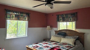 A bedroom with a bed, a ceiling fan, two windows with curtains, and a nightstand.