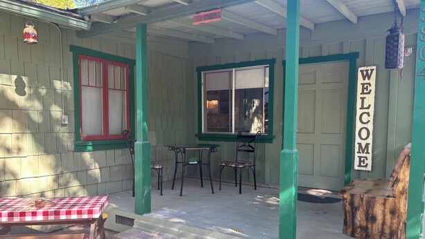 A porch with a red and white checkered table, two chairs, and a wooden bench. A 'WELCOME' sign is visible on the wall.