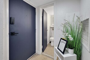 Um corredor com uma porta azul escura, uma parede branca com um quadro de boas-vindas e um vaso de plantas com flores brancas.