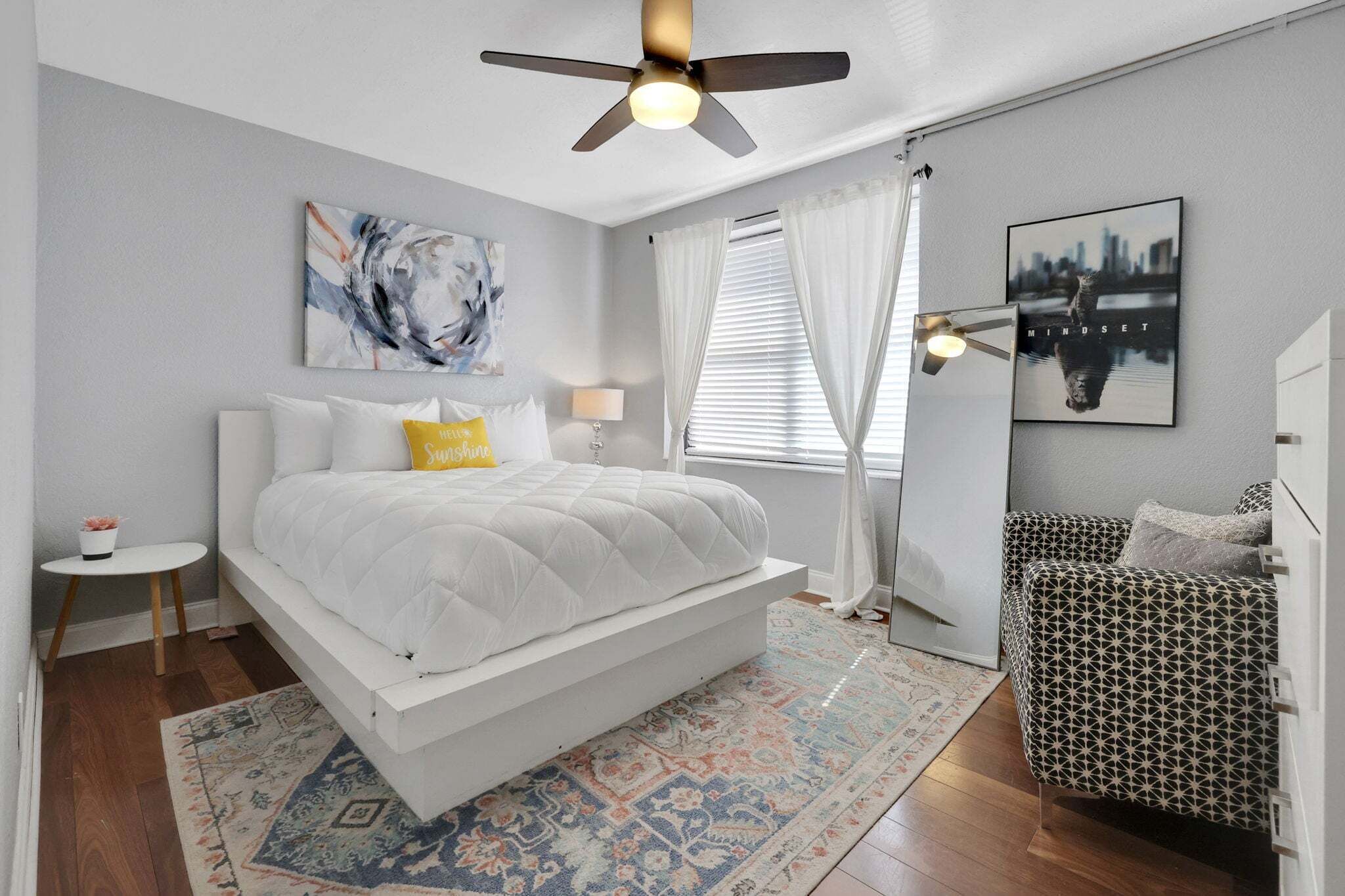 Um quarto com cama, mesa de cabeceira, luminária de chão, janela com cortinas, pintura de paisagem urbana e ventilador de teto.