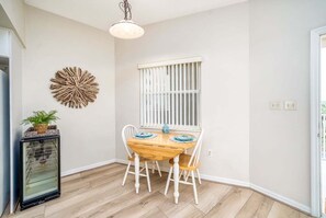 Ein Essbereich mit Holztisch und -stühlen, einer Zierdeko-Wandverkleidung, einem Fenster mit Jalousien und einem kleinen Kühlschrank.