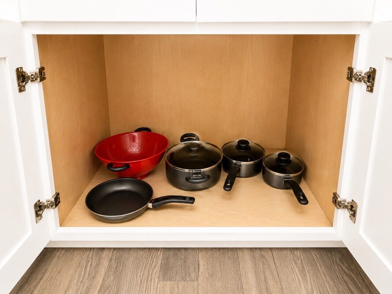 A kitchen cabinet with a wooden interior, containing a red colander, a black frying pan, and three black pots.
