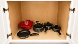 A kitchen cabinet with a wooden interior, containing a red colander, a black frying pan, and three black pots.