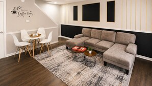 Ein modernes Wohnzimmer mit einer Eckcouch, einem Essbereich mit weißen Stühlen, einem Couchtisch und einem Deckenventilator.