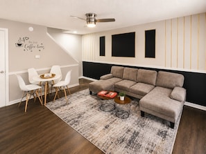 Un salon moderne avec un canapé d’angle, un coin repas avec des chaises blanches, une table basse et un ventilateur de plafond.