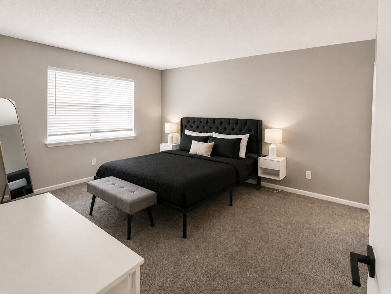 A bedroom with a bed, bedside tables, a bench, a mirror, and a window with blinds.