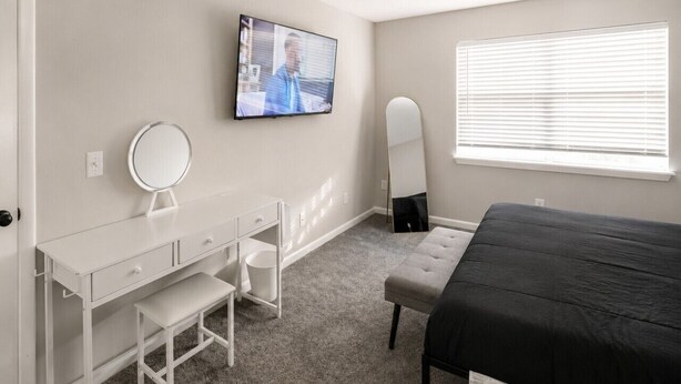 A bedroom with a bed, a desk with a mirror, a television, and a window with blinds.
