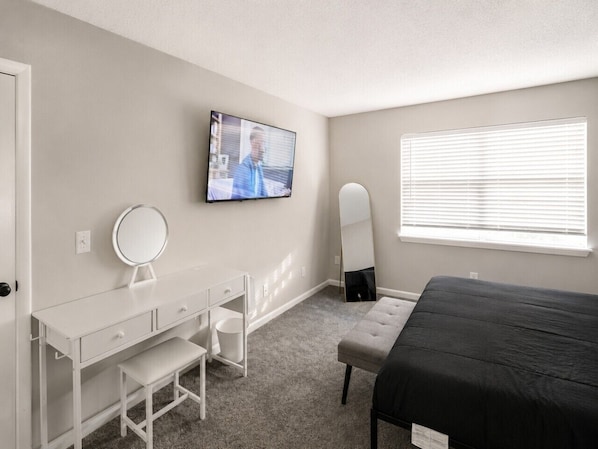 A bedroom with a bed, a desk with a mirror, a television, and a window with blinds.