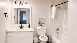 Een badkamer met een witte wastafel, toilet en bad, een spiegel boven de wastafel en een schilderij aan de muur.
