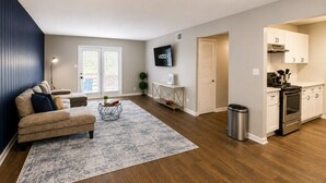 A modern living room with a sofa, a coffee table, and a rug. There is a kitchen area with cabinets and a stove.