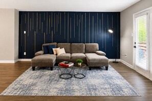 A sectional sofa with a geometric wall design, a patterned rug, and a small side table with a plant. - Modern Hoover Retreat | King + Queen (Hoover)