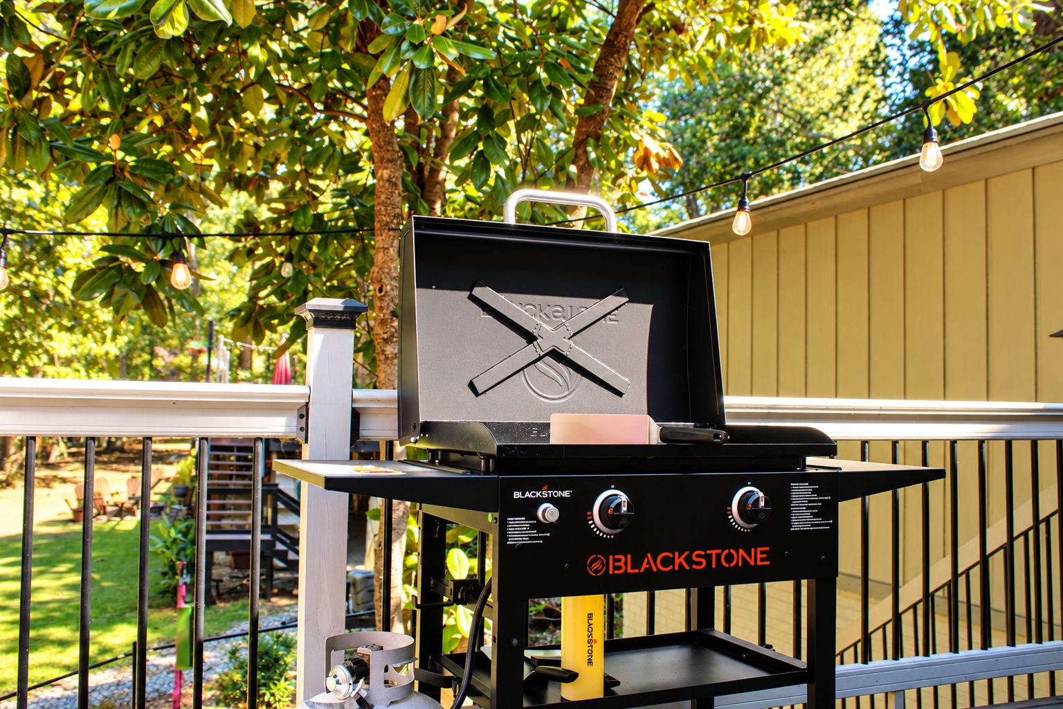 A BLACKSTONE grill with a lid open, placed on a balcony with a railing, surrounded by greenery and a house in the background.