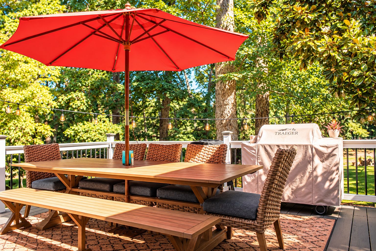 An outdoor dining area with a wooden table, wicker chairs, a red umbrella, and a grill station.