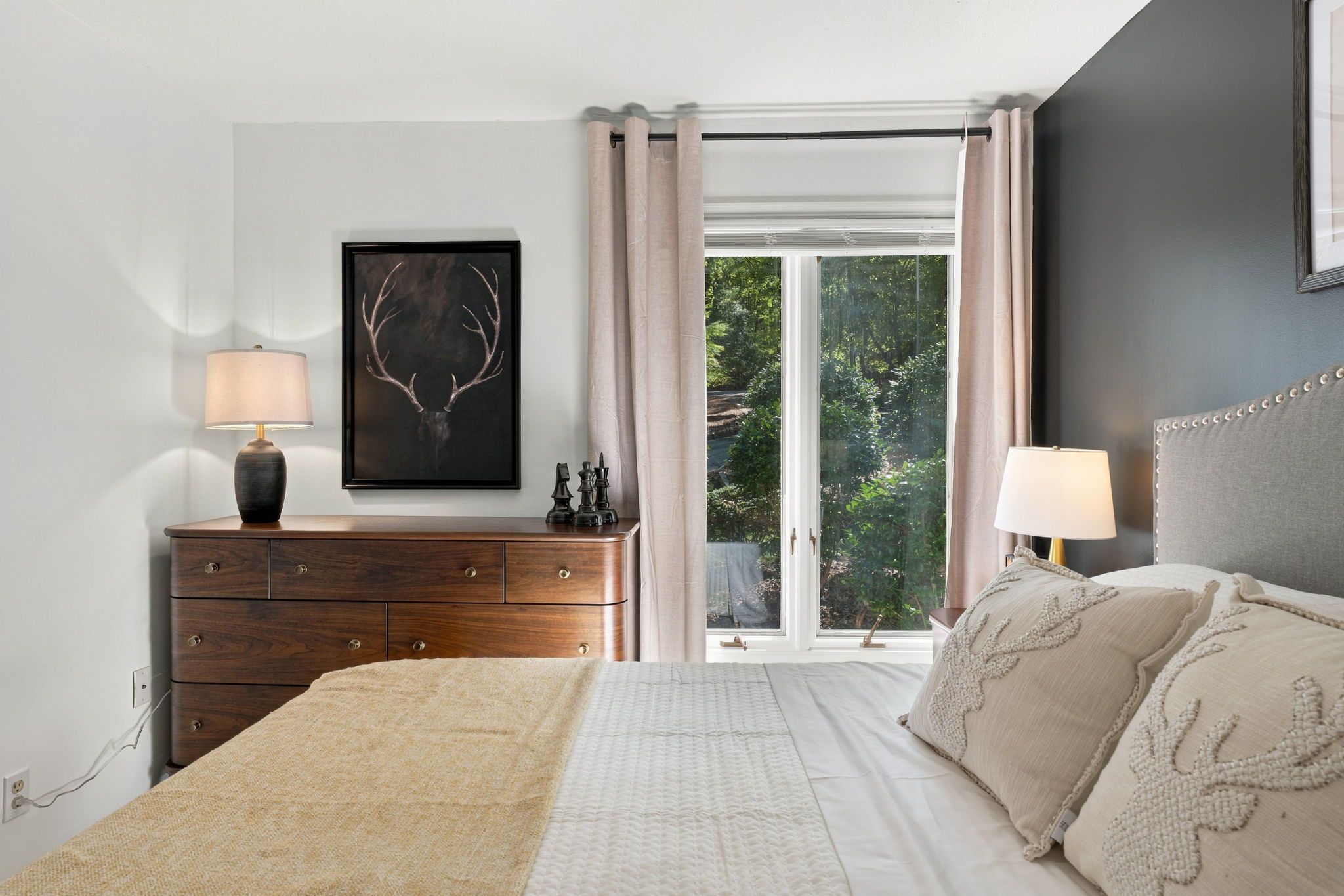 A bedroom with a bed, two bedside tables, a dresser, a window with curtains, and a framed deer head artwork.
