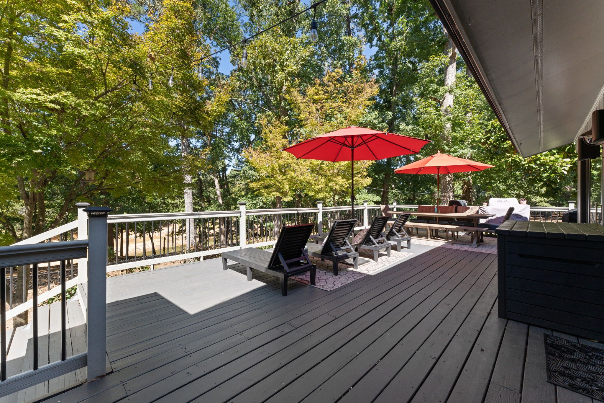 Terraza de madera con mobiliario exterior, incluyendo sillones y una mesa, bajo una zona cubierta con sombrillas rojas.