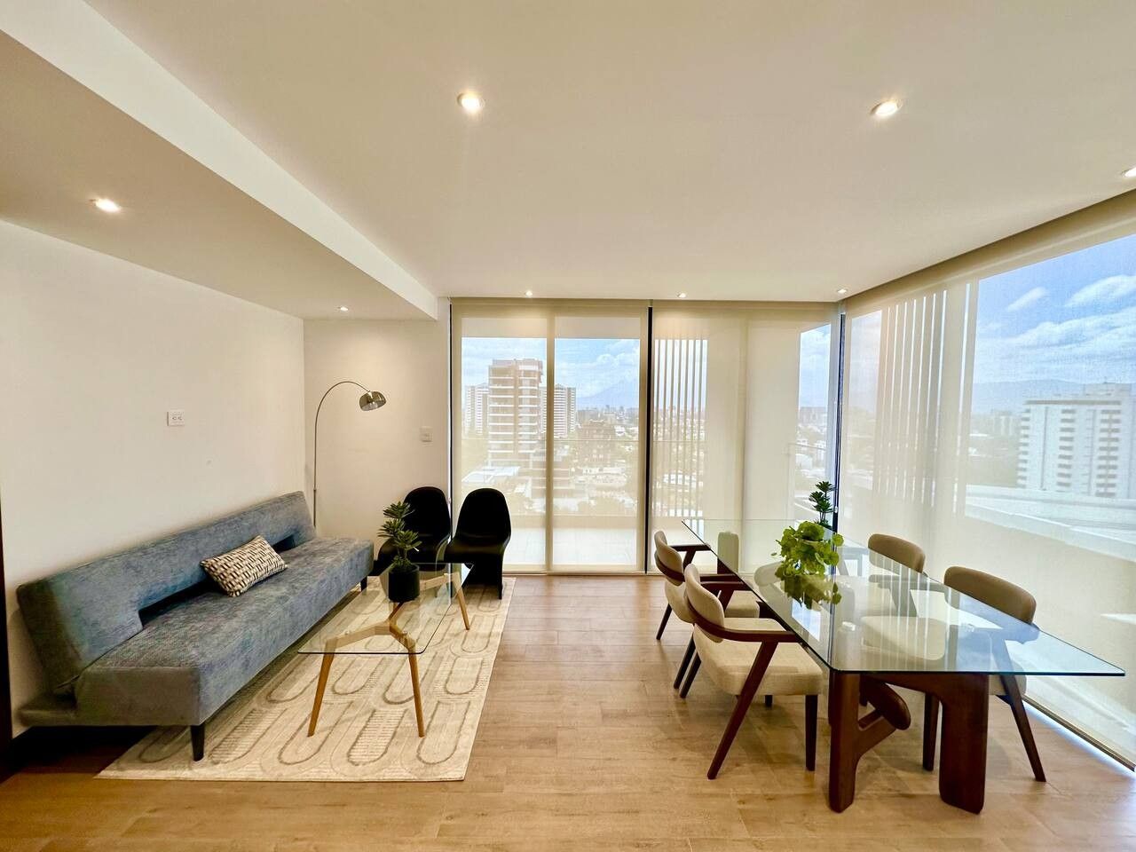 A modern living room with a sofa, chairs, a glass dining table, and a view through large windows.