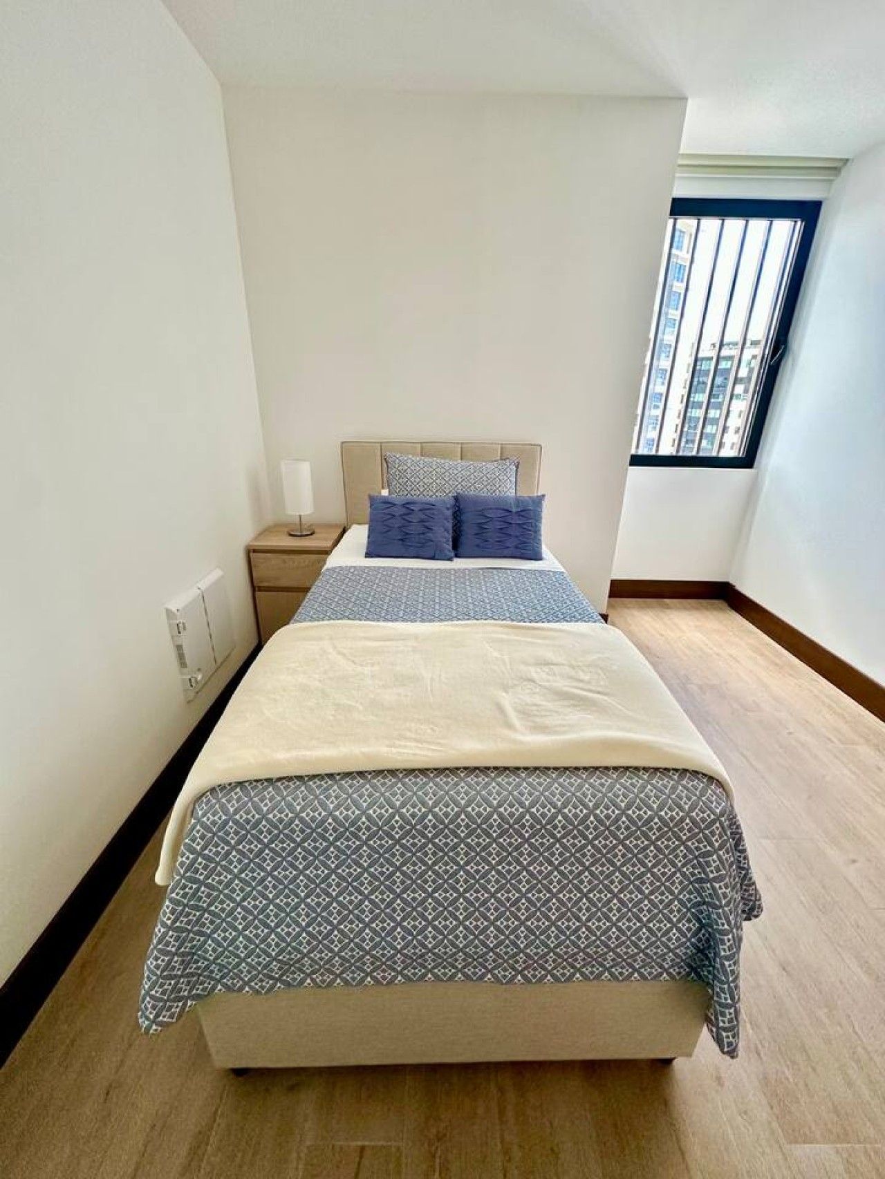Une chambre à coucher avec un lit, une table de chevet, une fenêtre et une vue sur des immeubles.