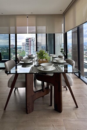 A modern dining area with a glass table, wooden chairs, and a city view. - New Luxury Condo 2BR/2BA | Zone 15 | Near Cayalá! (Guatemala City)