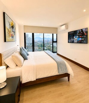 A modern bedroom with a large bed, a TV mounted on the wall, and a city view through the windows. - New Luxury Condo 2BR/2BA | Zone 15 | Near Cayalá! (Guatemala City)