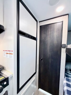 A compact room with a dark wooden door, a white wall with a safety notice, and a small bed with a checkered blanket. - Downtown New RV | 2 Blocks to Momentary & Trails (Bentonville)