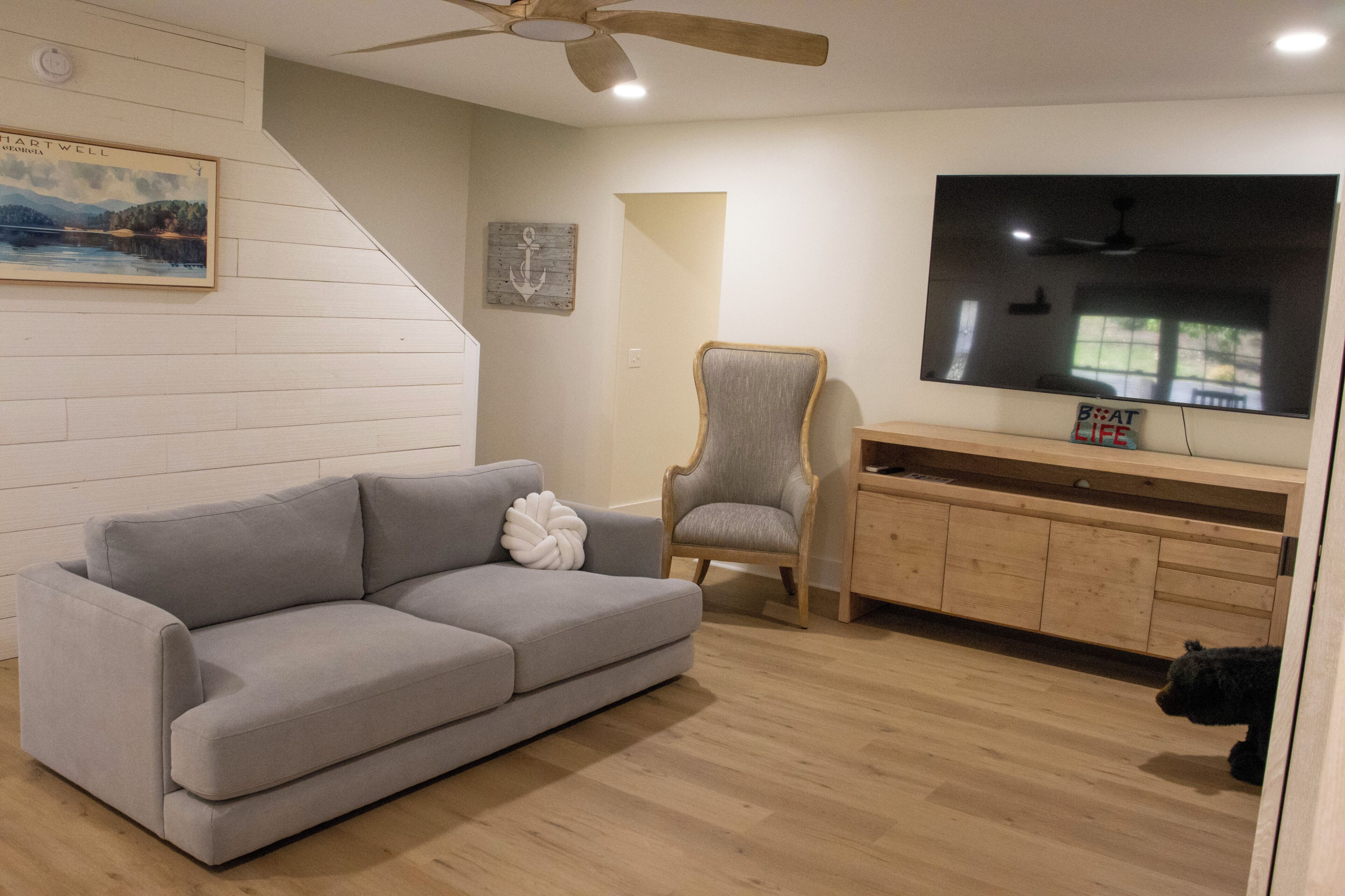 A living room with a grey sofa, a wooden chair, a TV on a wooden stand, and a ceiling fan.