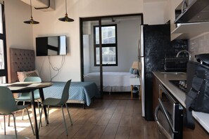 A compact room with a bed, a dining table, chairs, a TV mounted on the wall, a kitchen area with a stove and oven, and a window with a view. - EON Zone 10: Luxury, views, and comfort all in one place. (Ciudad de Guatemala)