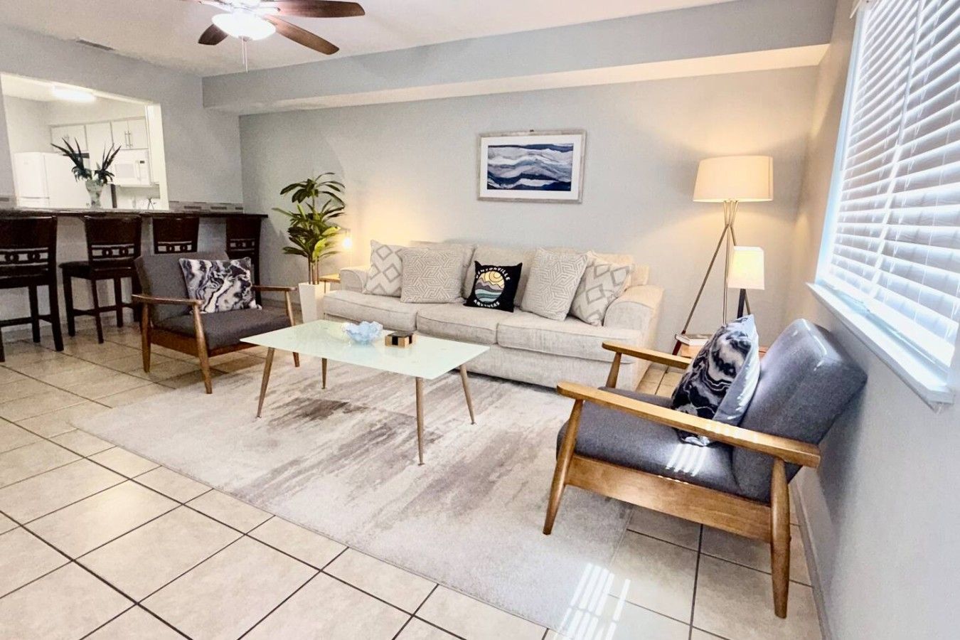 A living room with a sofa, two armchairs, a coffee table, a dining area with chairs, a ceiling fan, a painting, and a window with blinds.