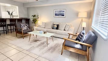 A living room with a sofa, two armchairs, a coffee table, a dining area with chairs, a ceiling fan, a painting, and a window with blinds.