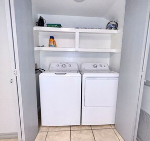 Une buanderie équipée de deux machines à laver et deux sèche-linge superposés, d’une étagère contenant des produits de lessive et d’une porte donnant sur une autre pièce.