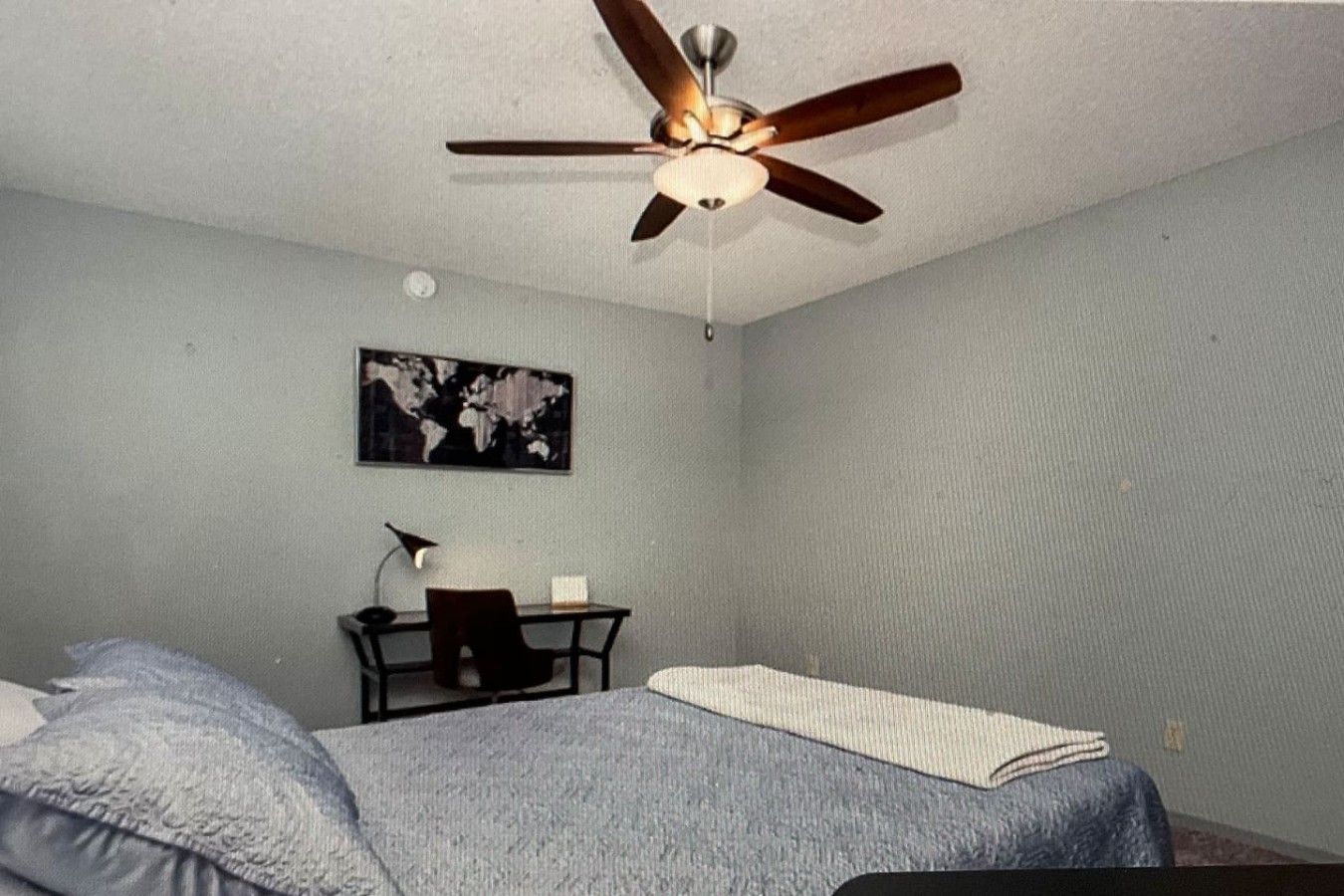 A bedroom with a bed, a desk, a chair, a ceiling fan, and a world map on the wall.