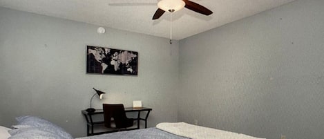 A bedroom with a bed, a desk, a chair, a ceiling fan, and a world map on the wall.