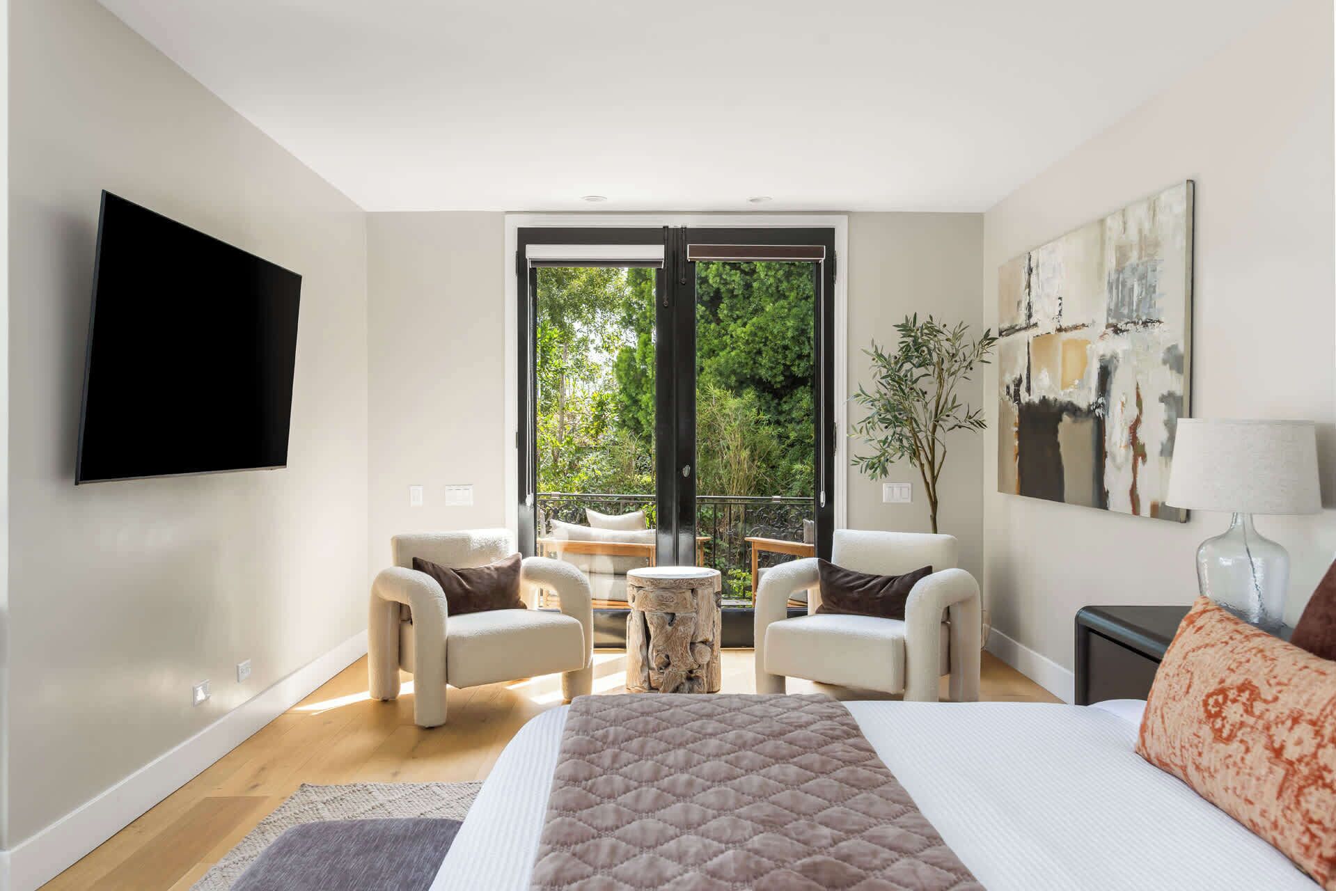 Ein Schlafzimmer mit einem Bett, zwei Sesseln, einem kleinen runden Tisch, einem Fernseher, einem Gemälde und Blick ins Grüne durch eine Schiebetür.